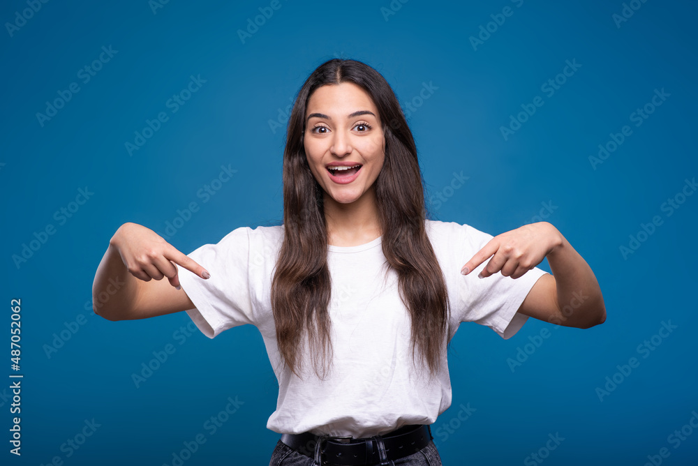Young and attractive caucasian or arab brunette girl in white t-shirt pointing fingers down at copy space for advertising isolated on blue studio background. Young and attractive caucasian or arab brunette girl in white t-shirt pointing fingers down at copy space for advertising isolated on blue studio background.