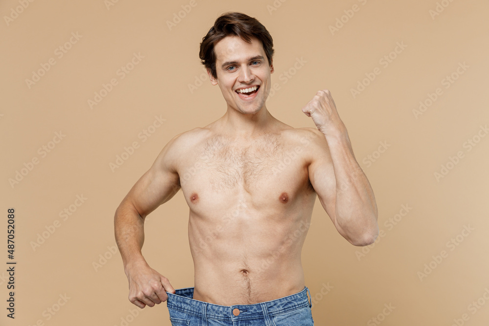 Fototapeta premium Shirtless young man 20s with naked torso showing loose pants do winner gesture isolated on pastel pastel beige background Body care healthcare fitness sport bodybuilding diet loosing weight concept