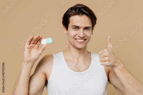 Happy young man 20s perfect skin wear undershirt hold contact lenses case container show thumb up isolated on pastel pastel beige background studio. Skin care healthcare cosmetic procedures concept.