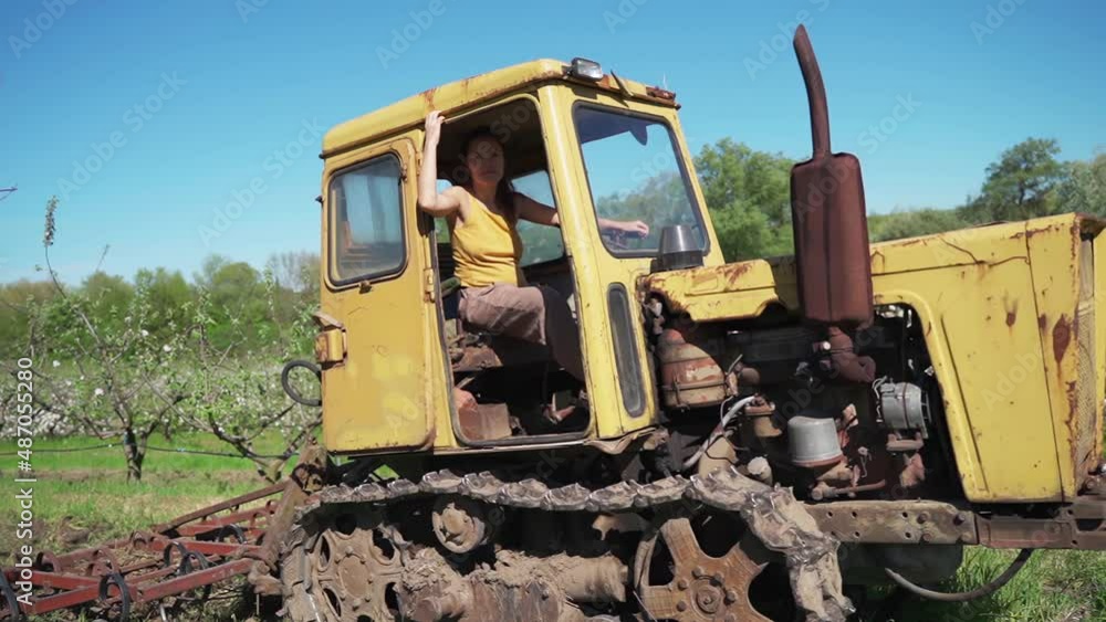 Old Tractor in the Field. Farmer in Old-fashioned tractor sowing crops at field. Woman driving small tractor in field and waving hand Old Tractor in the Field. Farmer in Old-fashioned tractor sowing crops at field. Woman driving small tractor in field and waving hand