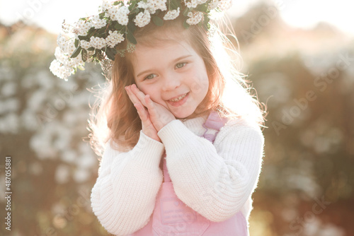 Smiling cute little child girl 3-4 year old wear floral wreath posing over white flowers at background. Springtime. Childhood. Happy baby girl with flower headband over blooming bushes in garden. © morrowlight