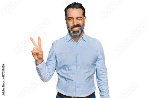 Middle aged man with beard wearing business shirt smiling with happy face winking at the camera doing victory sign with fingers. number two.