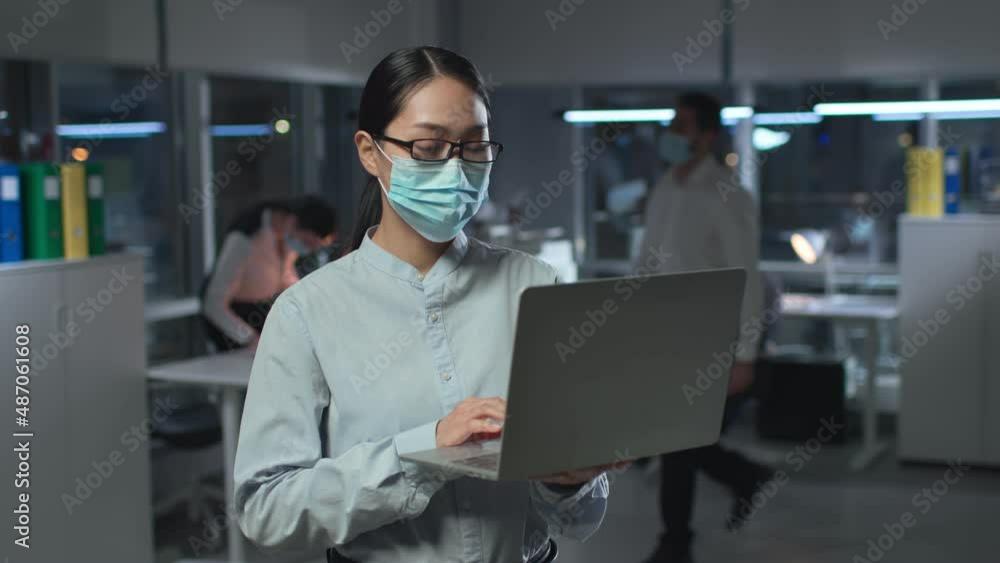 Portrait of asian businesswoman in face mask typing on laptop standing in office Portrait of asian businesswoman in face mask typing on laptop standing in office