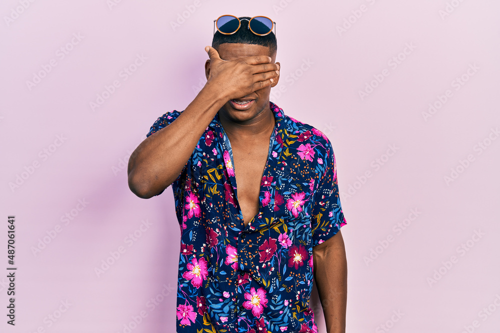 Young black man wearing hawaiian shirt and sunglasses smiling and laughing with hand on face covering eyes for surprise. blind concept. Young black man wearing hawaiian shirt and sunglasses smiling and laughing with hand on face covering eyes for surprise. blind concept.