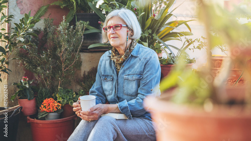 Thoughtful older woman, sitting between plants, holding cup