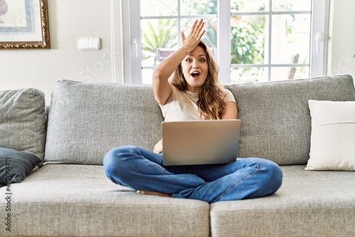 Beautiful young brunette woman sitting on the sofa using computer laptop at home surprised with hand on head for mistake, remember error. forgot, bad memory concept. photo