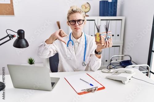 Young doctor woman holding model of human anatomical skin and hair at the clinic with angry face  negative sign showing dislike with thumbs down  rejection concept