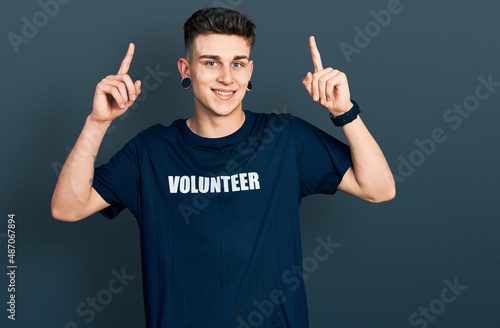 Young caucasian boy with ears dilation wearing volunteer t shirt smiling amazed and surprised and pointing up with fingers and raised arms.