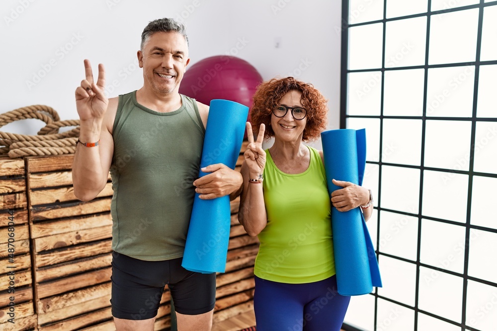 Middle age couple holding yoga mat smiling with happy face winking at the camera doing victory sign with fingers. number two.