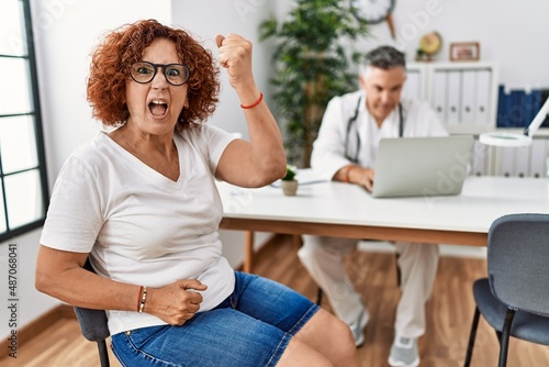 Senior woman sitting at doctor appointment angry and mad raising fist frustrated and furious while shouting with anger. rage and aggressive concept.