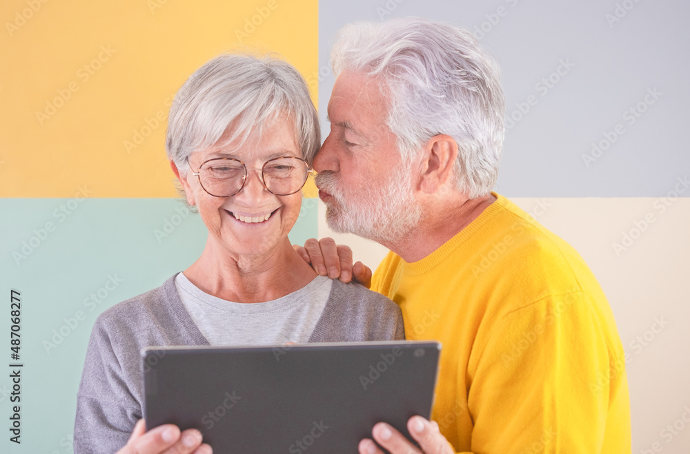 Portrait of smiling caucasian senior couple standing over isolated colorful background looking at same tablet. Elderly man kissing his wife. Retired man and woman using technology and social media Portrait of smiling caucasian senior couple standing over isolated colorful background looking at same tablet. Elderly man kissing his wife. Retired man and woman using technology and social media