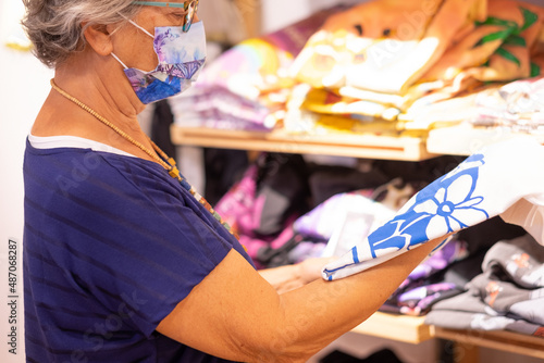 Coronavirus, Senior woman wearing protective face mask looking and choosing a t-shirt in shop retail. Mature elderly lady enjoying shopping in fashion store