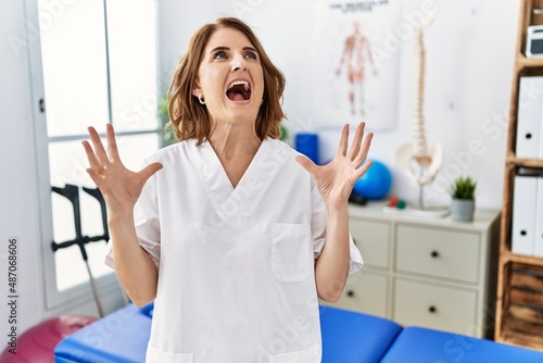Middle age physiotherapist woman working at pain recovery clinic crazy and mad shouting and yelling with aggressive expression and arms raised. frustration concept.