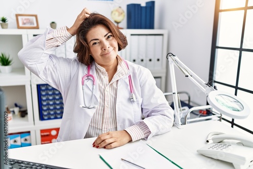 Middle age hispanic woman wearing doctor uniform and stethoscope at the clinic confuse and wonder about question. uncertain with doubt  thinking with hand on head. pensive concept.