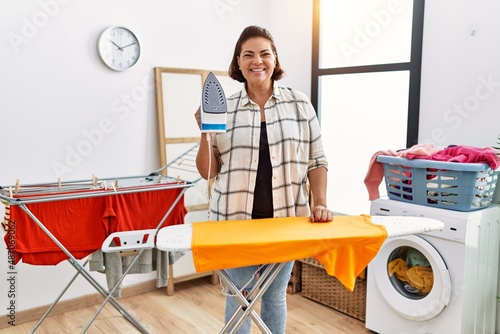Fotografía Middle age hispanic woman ironing clothes at home looking positive and happy sta