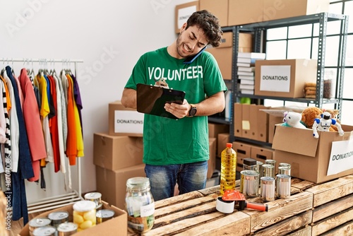 Young hispanic man wearing volunteer uniform writing on clipboard and talking on the smartphone at charity center