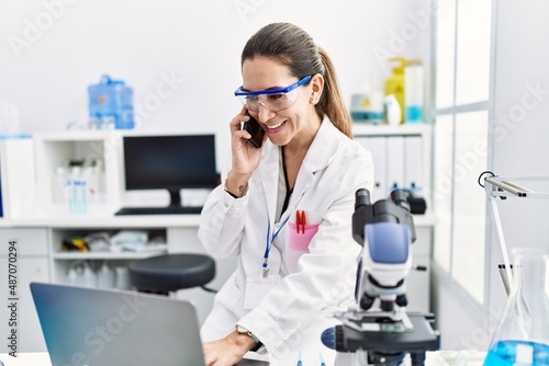 Young hispanic woman wearing scientist uniform talking on the smartphone at laboratory