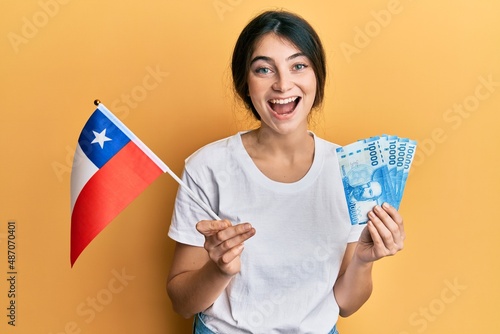 Young caucasian woman holding chile flag and chilean pesos banknotes smiling and laughing hard out loud because funny crazy joke. photo