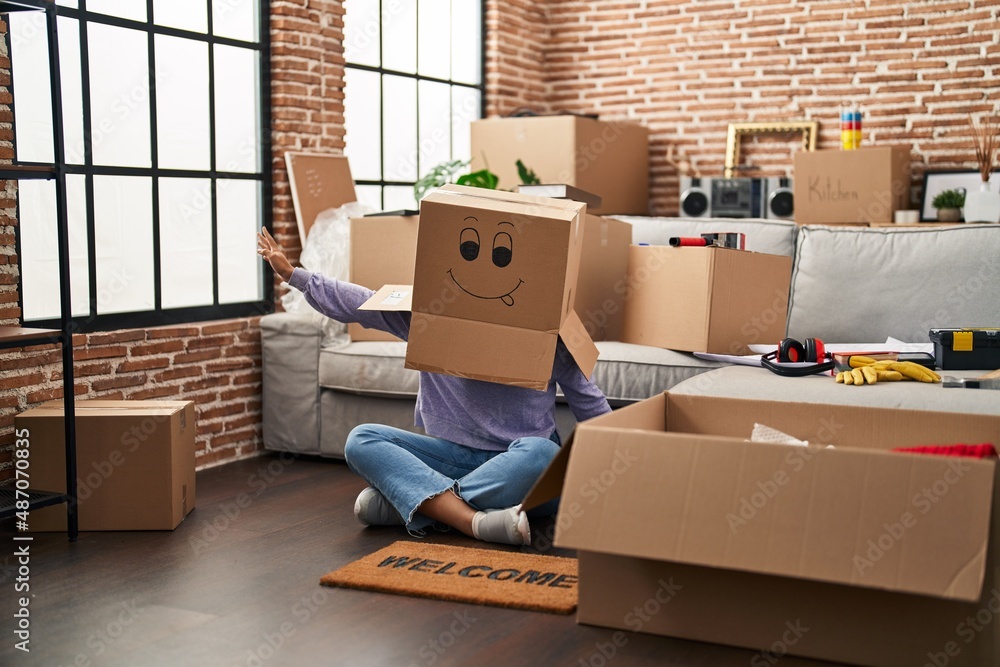 Fototapeta premium Young african american woman sitting on the floor with funny cardboard box on head at new home