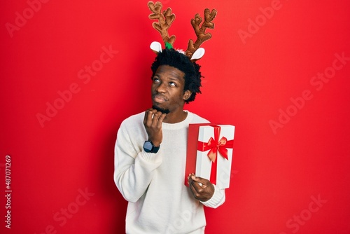 Young african american man wearing deer christmas hat holding gift with hand on chin thinking about question, pensive expression. smiling with thoughtful face. doubt concept.