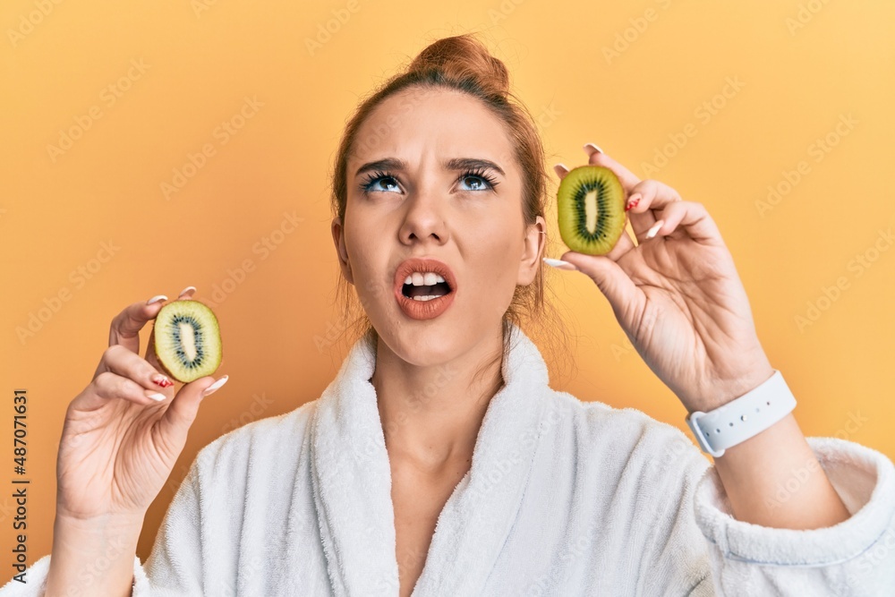 Young blonde woman wearing bathrobe holding fresh kiwi angry and mad screaming frustrated and furious, shouting with anger looking up. Young blonde woman wearing bathrobe holding fresh kiwi angry and mad screaming frustrated and furious, shouting with anger looking up.
