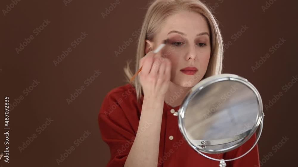 Beautiful blond hair woman in red shirt applying makeup in opposite of mirror Beautiful blond hair woman in red shirt applying makeup in opposite of mirror
