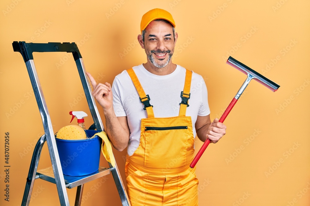 Handsome middle age man with grey hair wearing glass cleaner uniform and squeegee smiling happy pointing with hand and finger to the side Handsome middle age man with grey hair wearing glass cleaner uniform and squeegee smiling happy pointing with hand and finger to the side