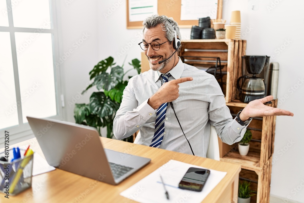 Middle age hispanic business man working at the office wearing operator headset amazed and smiling to the camera while presenting with hand and pointing with finger. Middle age hispanic business man working at the office wearing operator headset amazed and smiling to the camera while presenting with hand and pointing with finger.