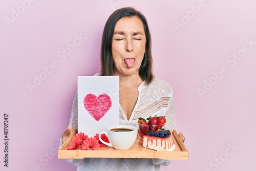 Middle age hispanic woman holding tray with breakfast food and heart draw sticking tongue out happy with funny expression.