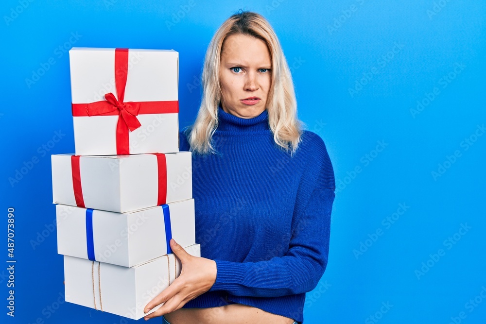 Beautiful caucasian blonde woman holding gifts clueless and confused expression. doubt concept. Beautiful caucasian blonde woman holding gifts clueless and confused expression. doubt concept.