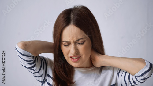 brunette woman with closed eyes touching neck and frowning from pain on grey background