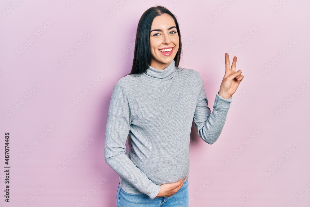 Beautiful woman with blue eyes expecting a baby, touching pregnant belly smiling looking to the camera showing fingers doing victory sign. number two. Beautiful woman with blue eyes expecting a baby, touching pregnant belly smiling looking to the camera showing fingers doing victory sign. number two.