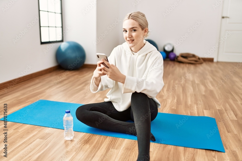 Young blonde girl relaxed using smartphone sitting on the floor at sport center. Young blonde girl relaxed using smartphone sitting on the floor at sport center.