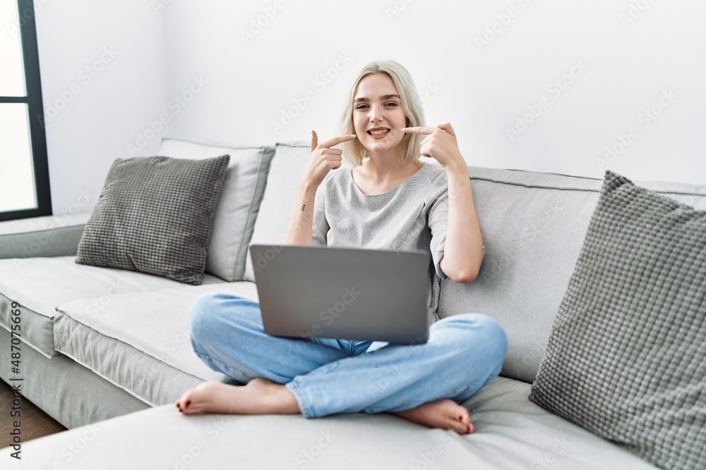 Fototapeta premium Young caucasian woman using laptop at home sitting on the sofa smiling cheerful showing and pointing with fingers teeth and mouth. dental health concept.