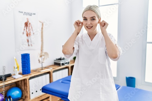 Young caucasian woman working at pain recovery clinic smiling pulling ears with fingers  funny gesture. audition problem