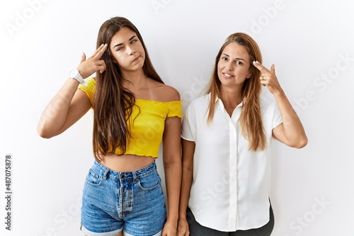 Mother and daughter together standing together over isolated background shooting and killing oneself pointing hand and fingers to head like gun, suicide gesture.