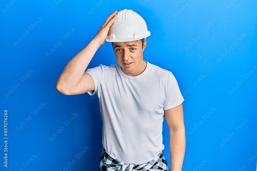 Handsome young man wearing builder uniform and hardhat surprised with hand on head for mistake, remember error. forgot, bad memory concept.