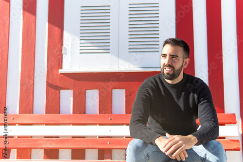 Young bearded boy sitting on a bench with a wall of red and white lines.