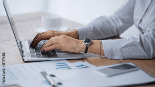 cropped view of woman typing on laptop near infographics on desk