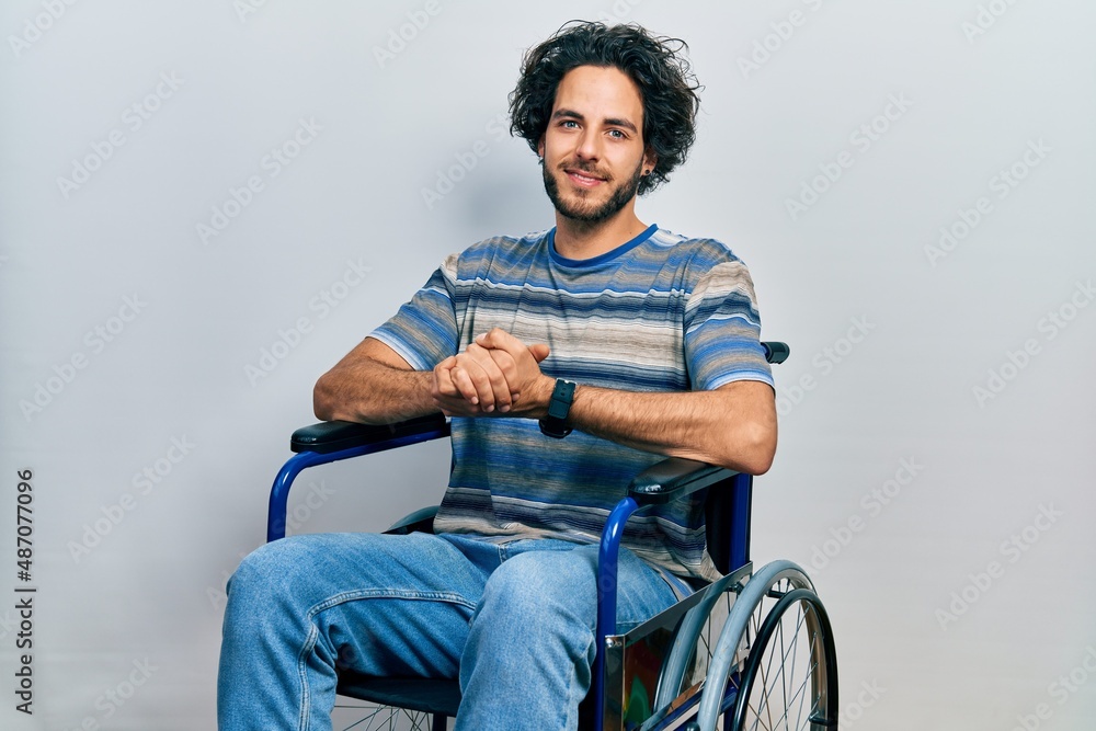 Handsome hispanic man sitting on wheelchair with hands together and crossed fingers smiling relaxed and cheerful. success and optimistic Handsome hispanic man sitting on wheelchair with hands together and crossed fingers smiling relaxed and cheerful. success and optimistic