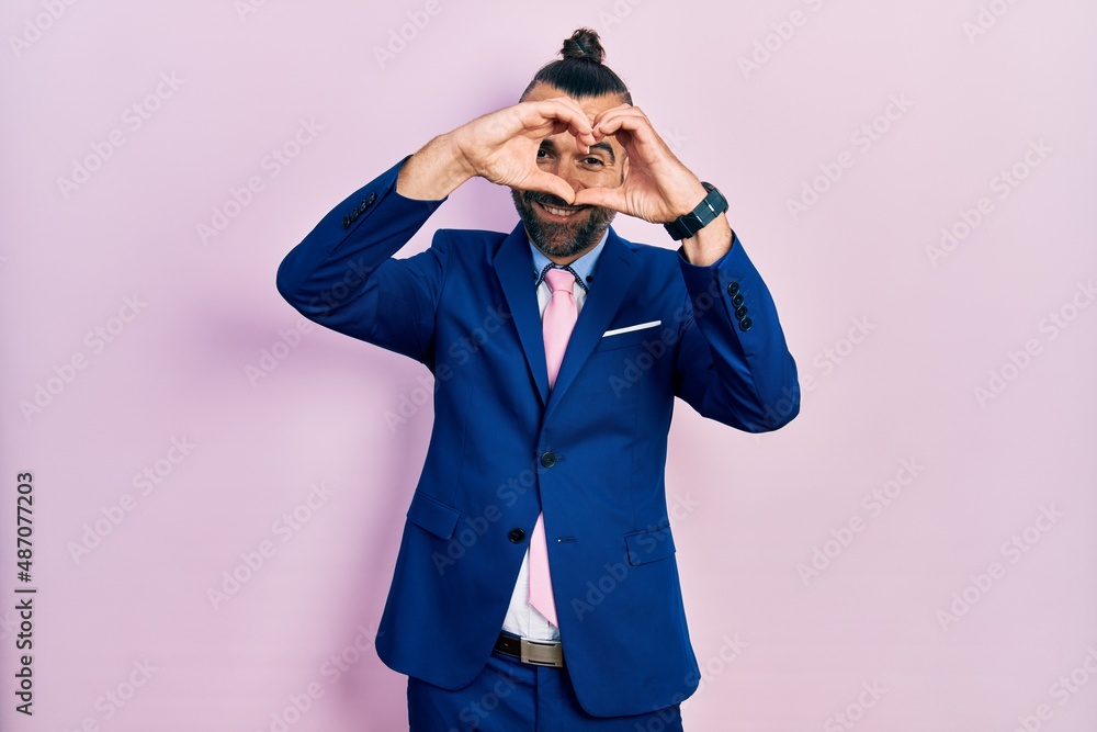 Young hispanic man wearing business suit and tie doing heart shape with hand and fingers smiling looking through sign Young hispanic man wearing business suit and tie doing heart shape with hand and fingers smiling looking through sign