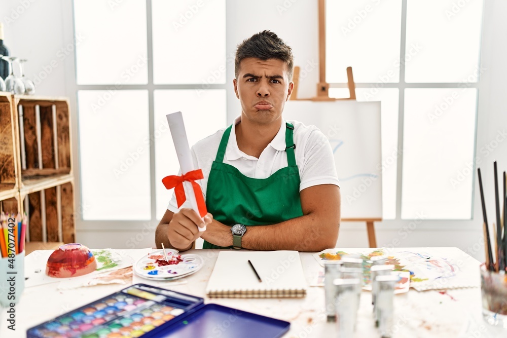 Young hispanic man at art studio holding degree depressed and worry for distress, crying angry and afraid. sad expression. Young hispanic man at art studio holding degree depressed and worry for distress, crying angry and afraid. sad expression.