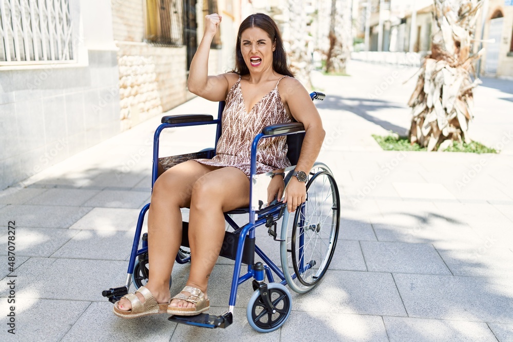 Young brunette woman sitting on wheelchair outdoors annoyed and frustrated shouting with anger, yelling crazy with anger and hand raised