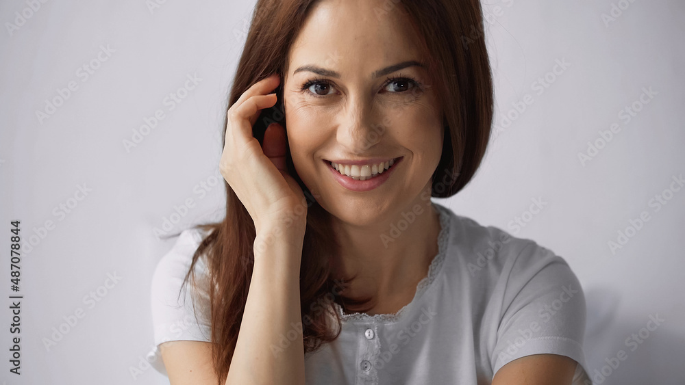 Fototapeta premium pleased brunette woman fixing hair and smiling at camera on grey background