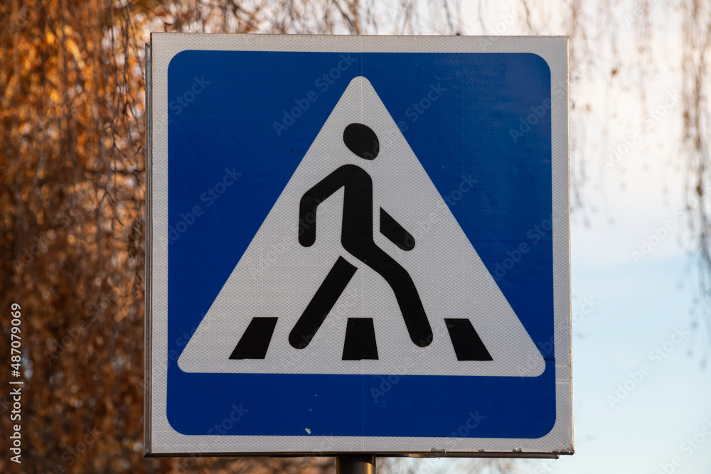 Traffic indicating sign, a blue crosswalk sign for pedestrians crossing the street or road on blur green trees and blue sky background in the city. No people.