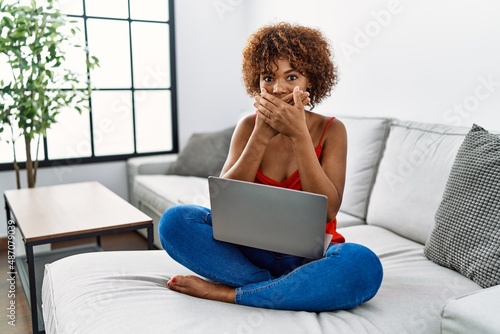Young african american woman sitting on the sofa at home using laptop shocked covering mouth with hands for mistake. secret concept.