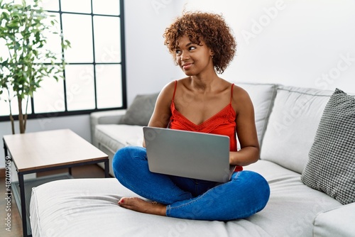 Young african american woman sitting on the sofa at home using laptop smiling looking to the side and staring away thinking.