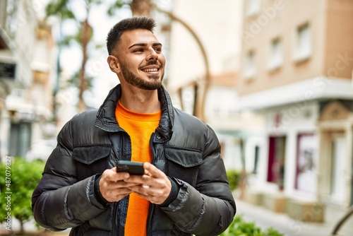 Handsome hispanic man with beard smiling happy and confident at the city wearing winter coat using smartphone
