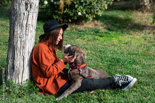 Una linda y alegre mujer caucásica con un sombrero está sentada en el parque, junto a su perro de la raza American Stafford Terrier