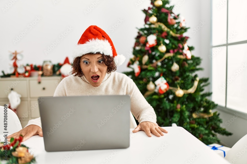 Middle age hispanic woman using laptop sitting by christmas tree scared and amazed with open mouth for surprise, disbelief face Middle age hispanic woman using laptop sitting by christmas tree scared and amazed with open mouth for surprise, disbelief face